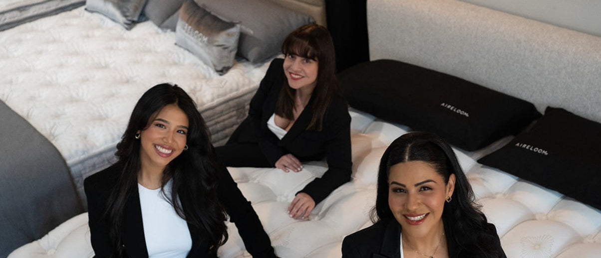 Three women in formal attire sitting on a bed with white bedding and black pillows.
