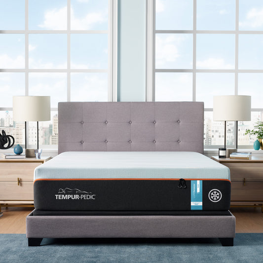 TEMPUR-LuxeBreeze Firm mattress in a stylish gray upholstered bed frame, positioned in a bright, modern bedroom with large windows and contemporary nightstands.