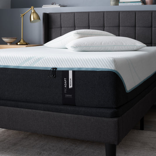 Tempur-Pedic ProAdapt Medium mattress in a modern bedroom setting, placed on a dark upholstered bed frame with white pillows and a gold lamp in the background.